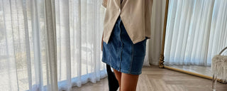 Woman wearing a beige top and denim skirt in a room with light curtains.