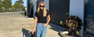 Woman in black top and blue jeans standing in front of a building with a plant nearby.