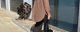 Woman in a brown sheer top holding a black bag on a sidewalk with a building in the background.