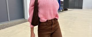 Woman wearing a pink knitted sweater and brown pants standing on a sidewalk.