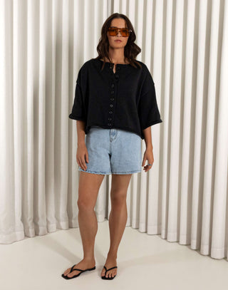 Woman wearing a black shirt and denim shorts standing against a white curtain background Black cotton button front top with relaxed shape and short sleeves