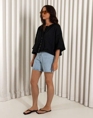 Woman wearing a black shirt and denim shorts standing against a white curtain background Black cotton button front top with relaxed shape and short sleeves