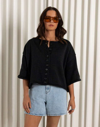 Woman wearing a black shirt and denim shorts standing against a white curtain background Black cotton button front top with relaxed shape and short sleeves