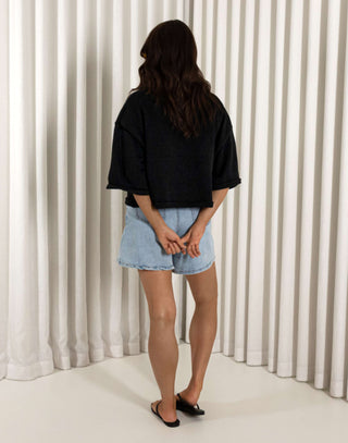 Woman wearing a black shirt and denim shorts standing against a white curtain background Black cotton button front top with relaxed shape and short sleeves