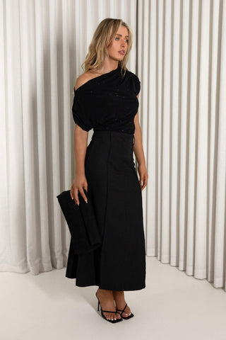 A woman models a black one-shoulder midi dress with a textured bodice and a high-waisted, flared skirt, holding a matching black clutch against a white pleated curtain backdrop.