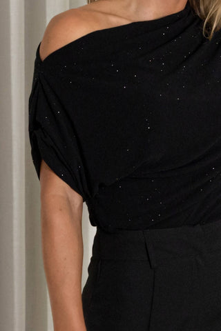 Close-up of a person wearing a black off-the-shoulder top with subtle silver sparkle details and black bottoms, cropped from the neck down against an indoor neutral background.