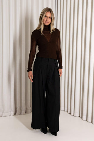 A woman modeling a dark brown sheer long-sleeve mock neck top paired with black wide-leg trousers, standing in front of a white pleated curtain backdrop.