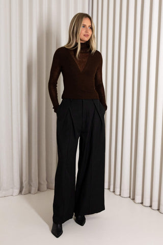 A woman modeling a dark brown mock-neck long-sleeve knit top paired with black wide-leg trousers, standing in front of a white pleated curtain backdrop.