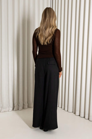 A woman viewed from behind wearing a dark brown long-sleeve knit top and black wide-leg trousers, standing in front of a white pleated curtain backdrop.