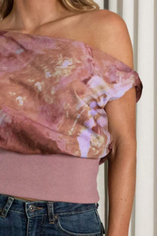 A close-up of a model wearing a dusty rose off-the-shoulder top with a large, draped mauve and pink abstract print overlay, paired with blue jeans.