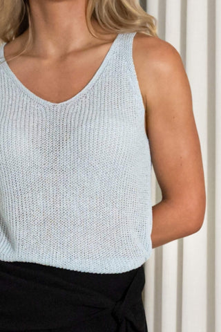 A close-up of a woman wearing a light blue-gray ribbed knit sleeveless tank top with a V-neck, paired with black bottoms, set against a neutral cream-colored curtain background.