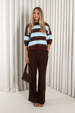 A woman stands in front of white pleated curtains wearing a fuzzy brown and light blue striped sweater, dark brown wide-leg trousers, and holding a brown handbag.