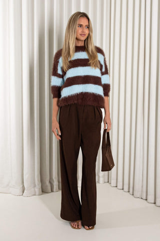 A woman stands in front of white pleated curtains wearing a fuzzy brown and light blue striped sweater, dark brown wide-leg trousers, and holding a brown handbag.