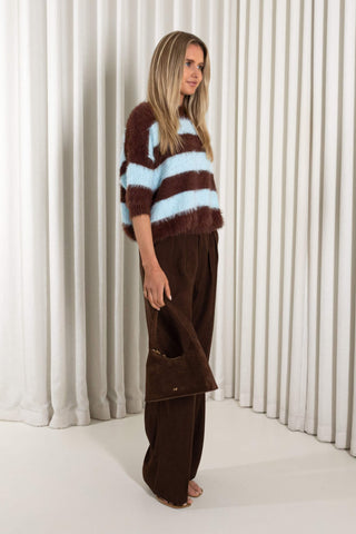 A woman stands in front of white pleated curtains wearing a fuzzy brown and light blue striped sweater, dark brown wide-leg trousers, and holding a brown handbag.