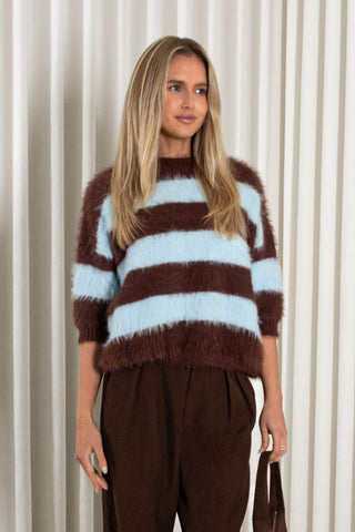 A woman stands in front of white pleated curtains wearing a fuzzy brown and light blue striped sweater, dark brown wide-leg trousers, and holding a brown handbag.