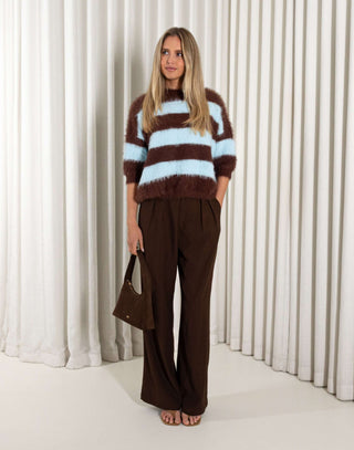 Woman wearing a brown and blue striped sweater with wide-leg pants against a white curtain background