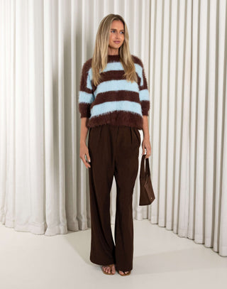 Woman wearing a brown and blue striped sweater with wide-leg pants against a white curtain background
