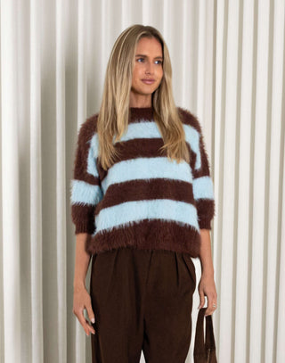 Woman wearing a brown and blue striped sweater with wide-leg pants against a white curtain background