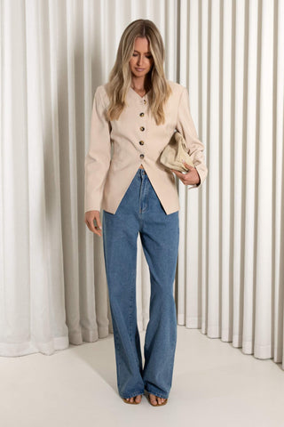 A woman stands in front of white pleated curtains wearing a light beige cropped jacket with dark buttons, blue wide-leg jeans, and holding a beige clutch.