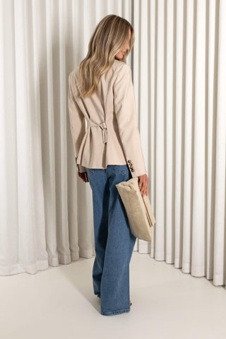 A woman stands in front of white pleated curtains wearing a light beige cropped jacket with dark buttons, blue wide-leg jeans, and holding a beige clutch.