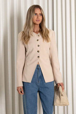 A woman stands in front of white pleated curtains wearing a light beige cropped jacket with dark buttons, blue wide-leg jeans, and holding a beige clutch.