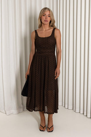 A woman modeling a sleeveless midi dress with a dark brown and black polka dot pattern, standing in front of a white curtain backdrop.
