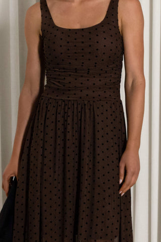 A woman wearing a sleeveless brown dress with a black polka dot pattern, featuring a square neckline and a gathered waist.