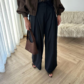 Person wearing a brown jacket and black wide-leg pants holding a brown handbag indoors.