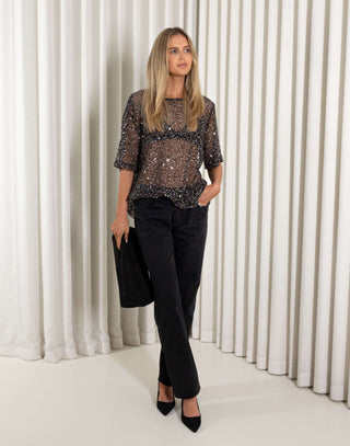Woman wearing a sequin top and black pants against a white curtain background