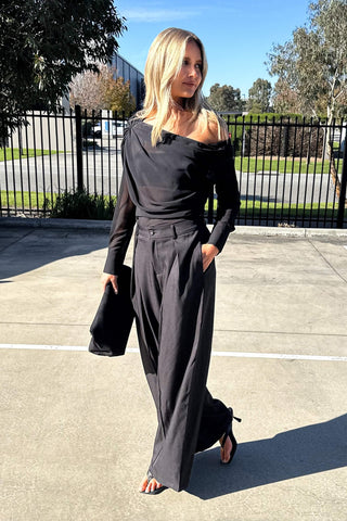A woman wearing a black off-the-shoulder long-sleeve top with wide-leg pants, holding a black clutch bag, standing outdoors on a paved surface.
