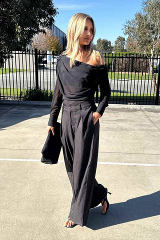A woman wearing a black off-the-shoulder long-sleeve top with wide-leg pants, holding a black clutch bag, standing outdoors on a paved surface.