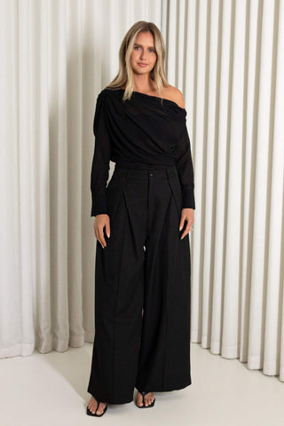 A woman modeling a black off-the-shoulder long-sleeve sheer top with wide-leg pants, standing against a white pleated curtain backdrop.