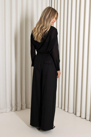 A woman wearing a black long-sleeve sheer top with high waist wide legs pants, photographed from the back against a white pleated curtain backdrop.