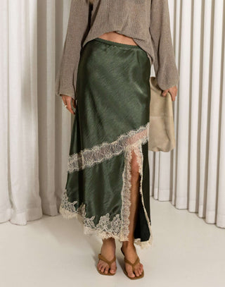 Woman wearing a green satin skirt with lace trim and a beige sweater against a white curtain background.
