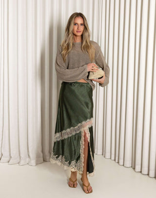 Woman wearing a green satin skirt with lace trim and a beige sweater against a white curtain background.