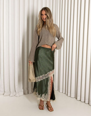 Woman wearing a green satin skirt with lace trim and a beige sweater against a white curtain background.