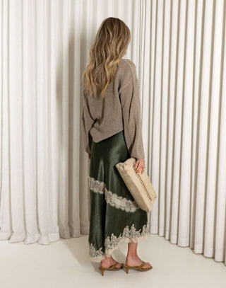 Woman wearing a green satin skirt with lace trim and a beige sweater against a white curtain background.