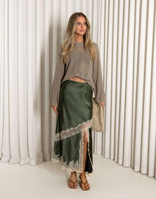 Woman wearing a green satin skirt with lace trim and a beige sweater against a white curtain background.