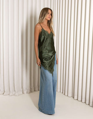 Woman wearing a green satin top and blue jeans against a white curtain background