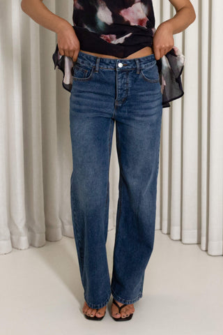 A person wearing blue wide-leg jeans, a dark floral top, and black sandals, standing in front of white curtains and holding the sides of the jeans to show the fit.
