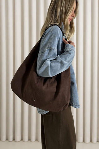 A woman wearing a light blue denim jacket and dark brown skirt carries a large, slouchy dark brown canvas tote bag over her shoulder, set against a background of vertical cream-colored drapes.