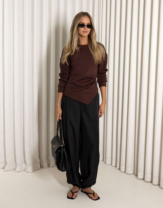 Woman wearing a brown top and black pants against a white curtain background
