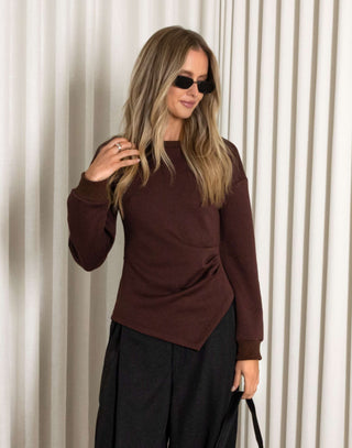 Woman wearing a brown top and black pants against a white curtain background