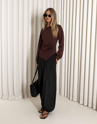 Woman wearing a brown top and black pants against a white curtain background
