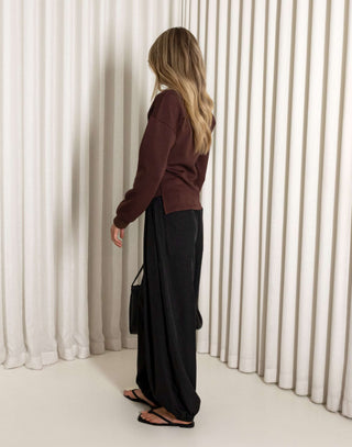 Woman wearing a brown top and black pants against a white curtain background