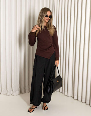 Woman wearing a brown top and black pants against a white curtain background