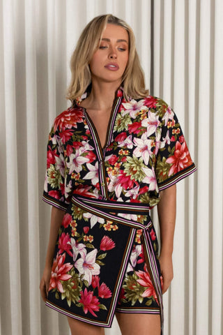A woman wearing a short-sleeve floral kimono-style mini dress with a wrap front and matching tie belt, standing against a white curtain backdrop.
