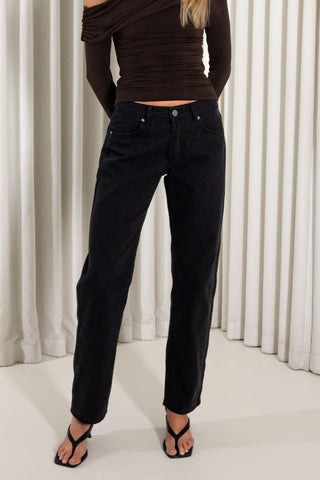 A model wearing black jeans, a dark brown off-the-shoulder long-sleeve top, and black strappy sandals, standing in front of a white curtain backdrop.