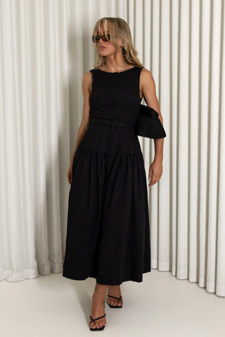 A woman modeling a sleeveless black midi dress with a structured bodice, gathered waist, and flared skirt, standing against a white pleated curtain backdrop.