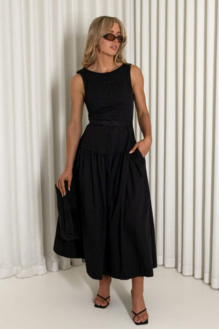 A woman modeling a sleeveless black midi dress with a buttoned bodice, matching belt, and side pockets, standing in front of white pleated curtains.